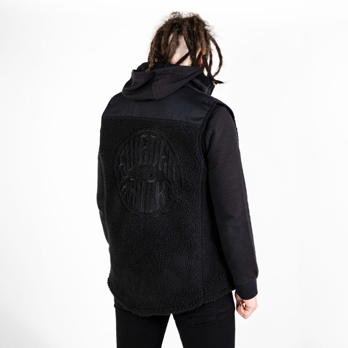 En person med dreadlocks, klädd i svarta byxor och luvtröja, bär Fleeceväst - en svart ärmlös väst med strukturerat material och präglade bokstäver på ryggen - vänd bort från kameran mot en vit bakgrund.