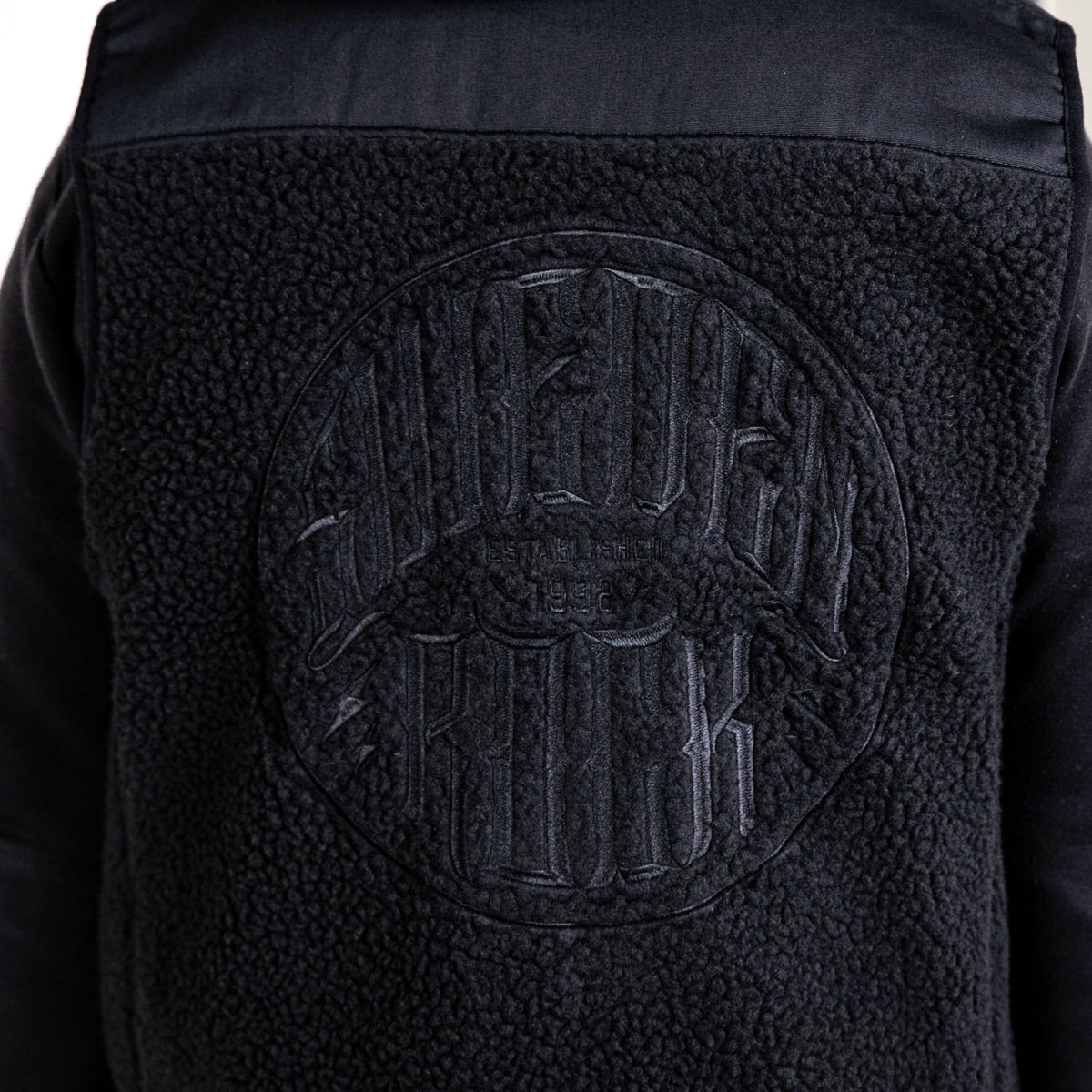 Fleecevästen har ett fleeceliknande svart material med slätt tyg på axlarna och en strukturerad cirkelformad patch på ryggen, prydd med upphöjt SUPER ROCK-broderi.