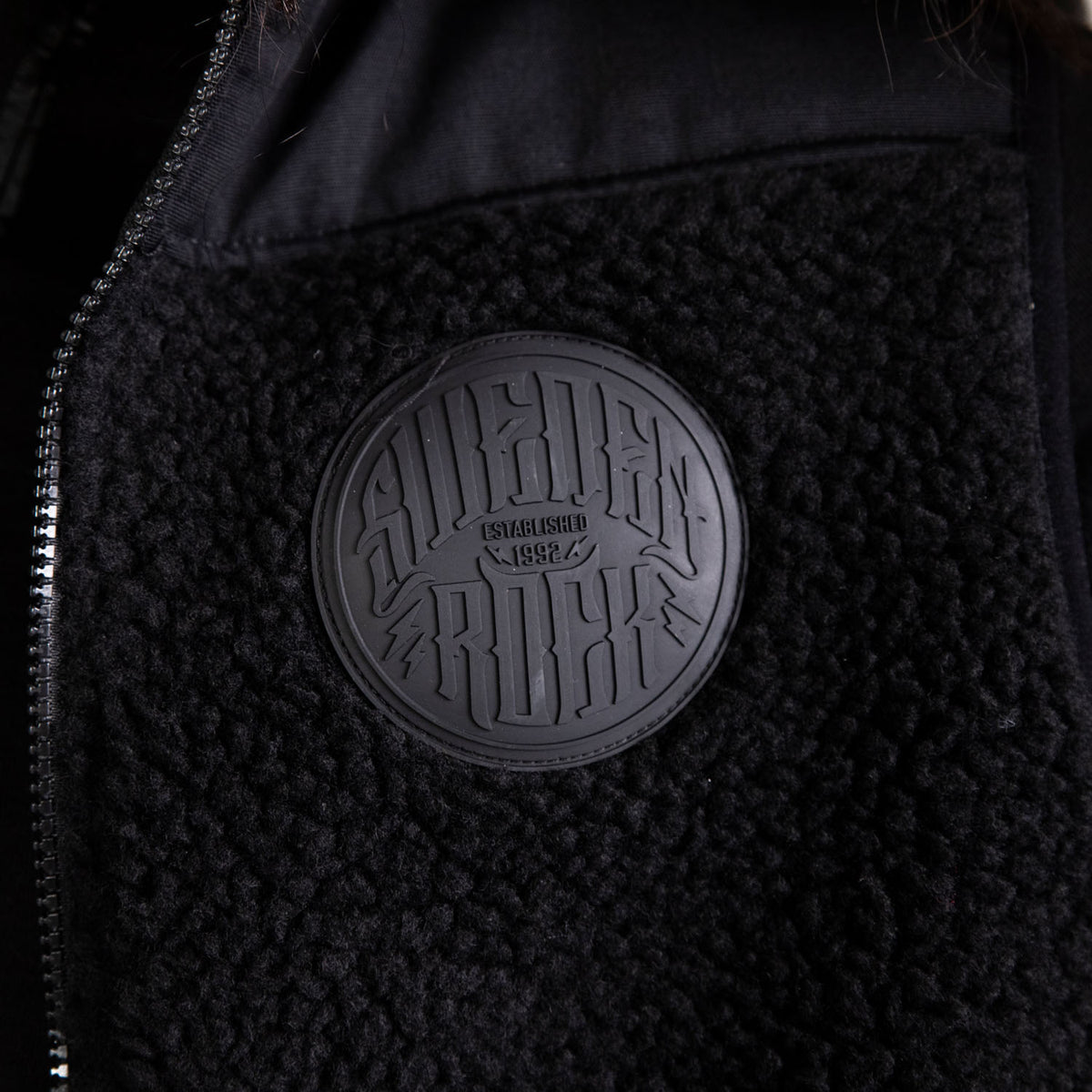 Närbild på Fleeceväst, en svart texturerad jacka med sherpafleece och en rund svart patch präglad med "Superdry Rock Established 1982".