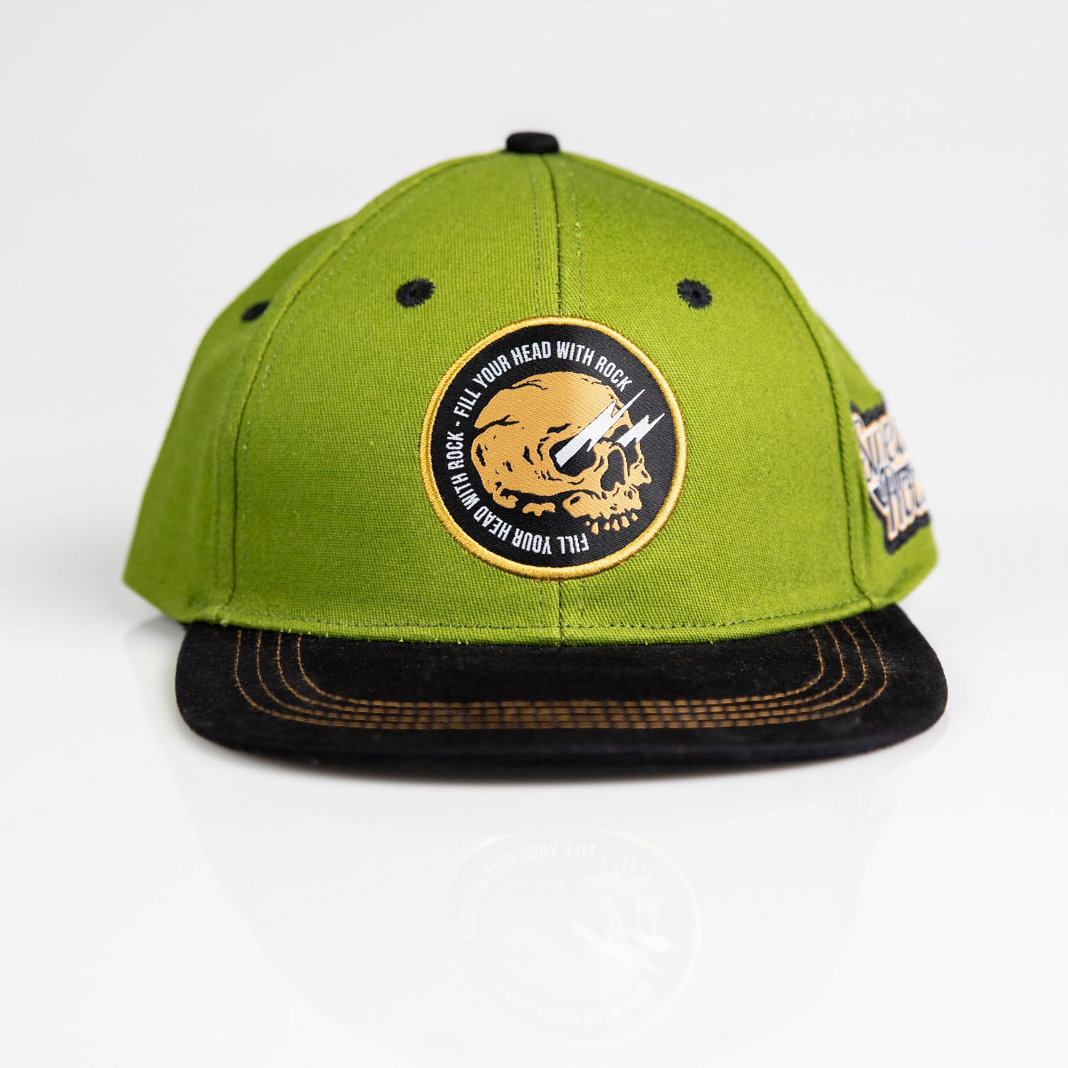 Keps Army/Black w patch är en grön snapback-keps med svart rak skärm, med en cirkulär dödskalle med stenar på framsidan och en annan patch på sidan, som visas mot en vit bakgrund.