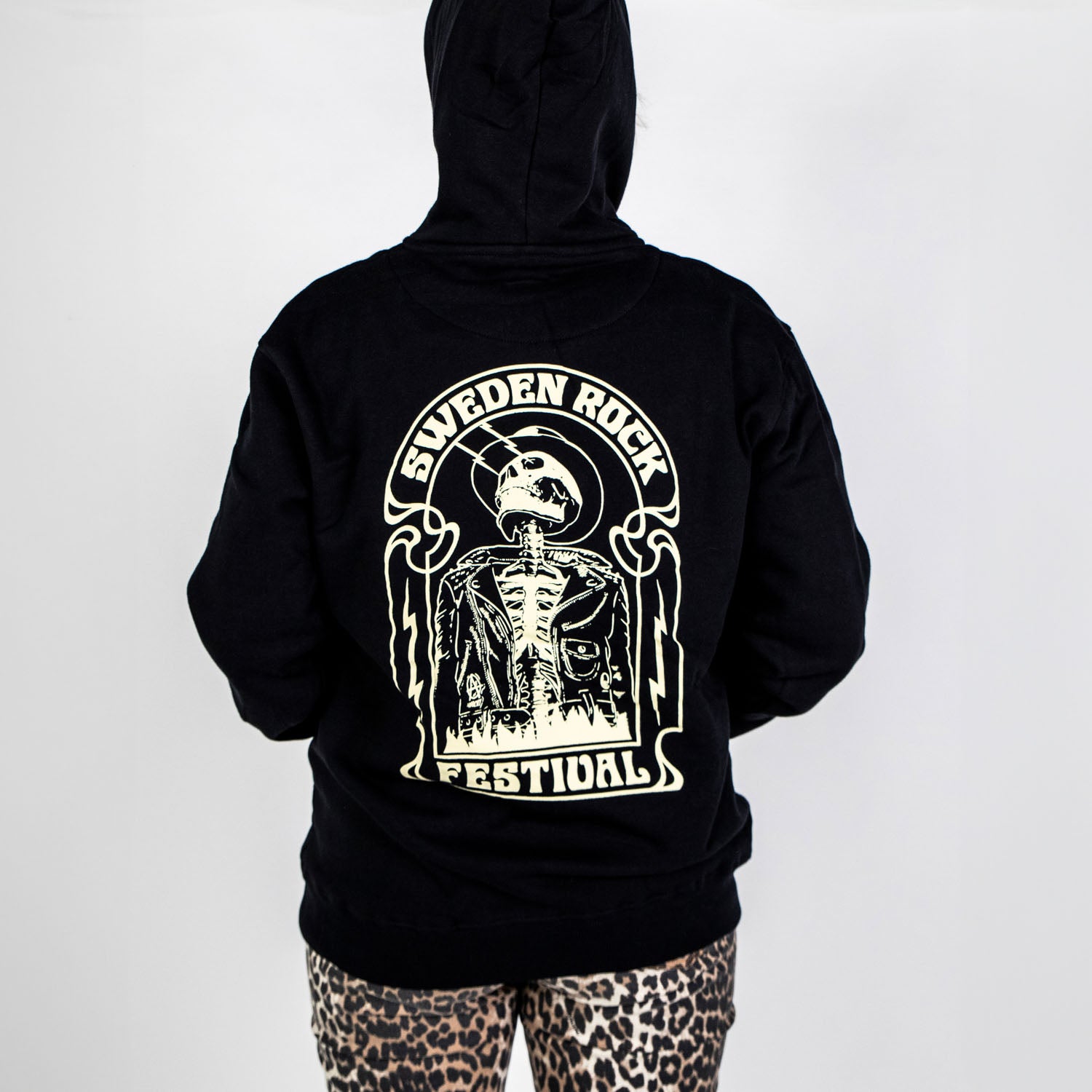 En person vänder sig bort från kameran och bär Zip Hoodie Skeleton Cowboy i svart polyester med Sweden Rock Festivals skelettgrafik på baksidan, tillsammans med leopardmönstrade byxor, allt mot en vit bakgrund.