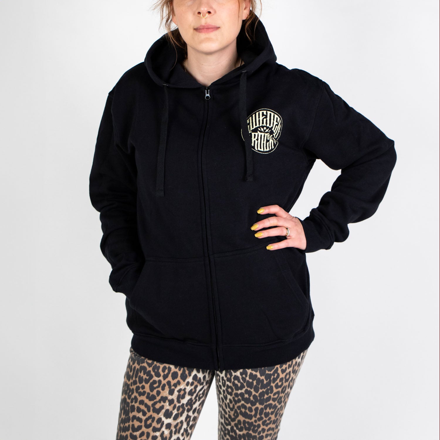 En person som bär Zip Hoodie Skeleton Cowboy - en svart huvtröja med dragkedja i ekologisk bomull med en rund vit logotyp på bröstet - och leggings med leopardmönster står med en hand på höften mot en vit bakgrund.
