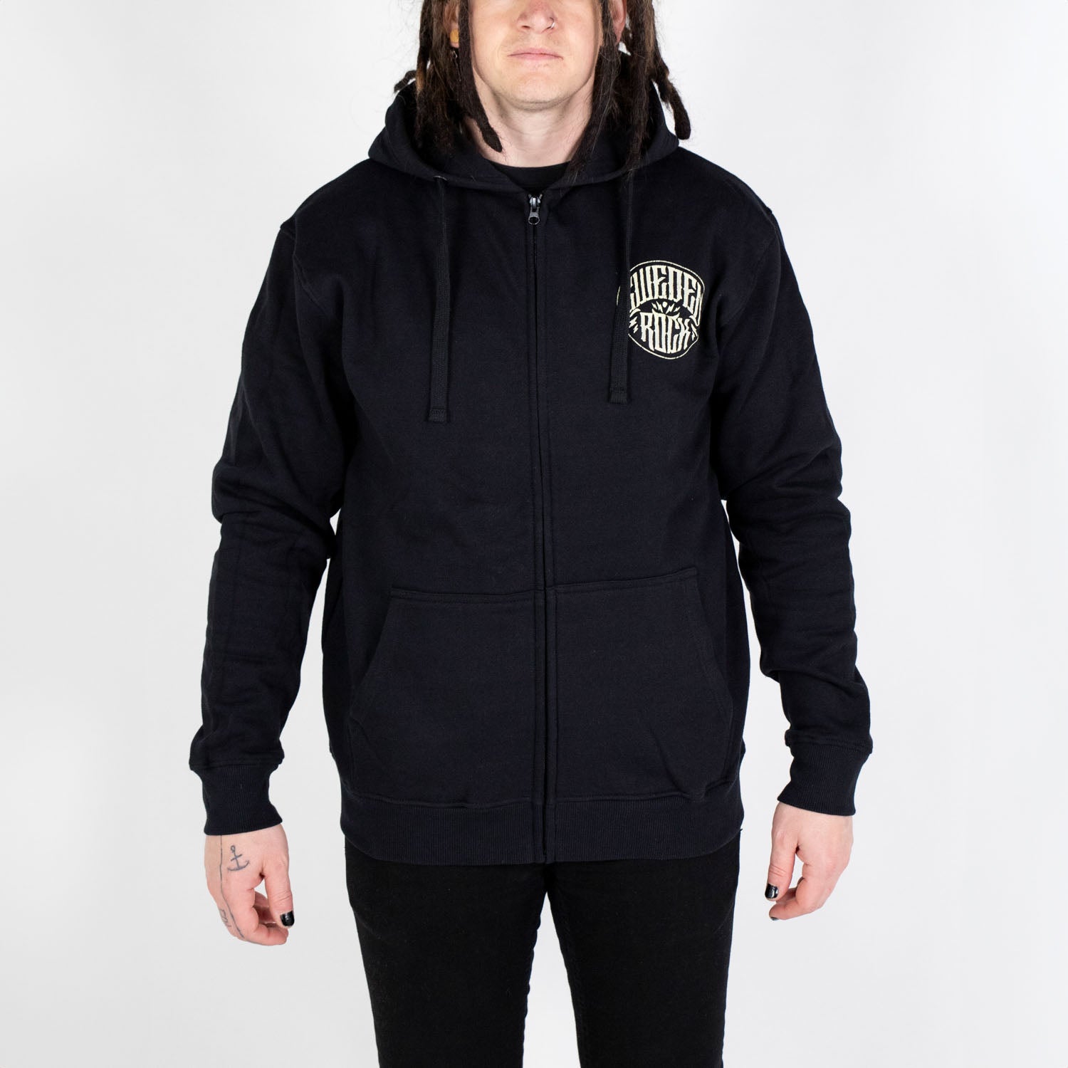 En person med långt brunt hår bär Zip Hoodie Skeleton Cowboy - svart, tillverkad av ekologisk bomull och polyester, med en vit cirkulär logotyp på vänster bröst och framfickor - stående mot en vit bakgrund, med armarna avslappnade vid sidan.