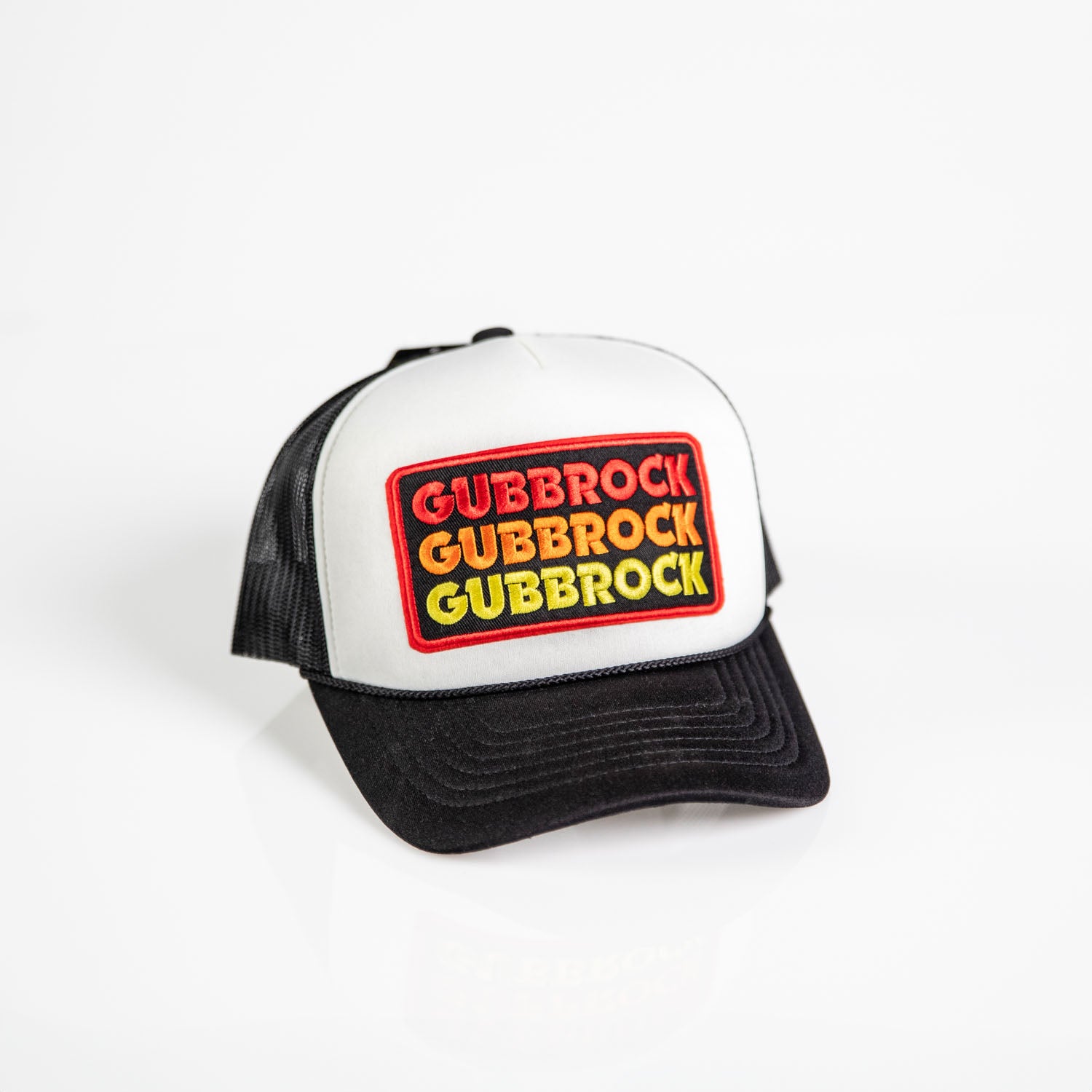 Keps Gubbrock är en svartvit truckerkeps med baksida i meshpolyester, snapback-stängning och en patch framtill som visar "GUBBROCK" tre gånger med röda, gula och gröna bokstäver. Visas på en vit bakgrund.