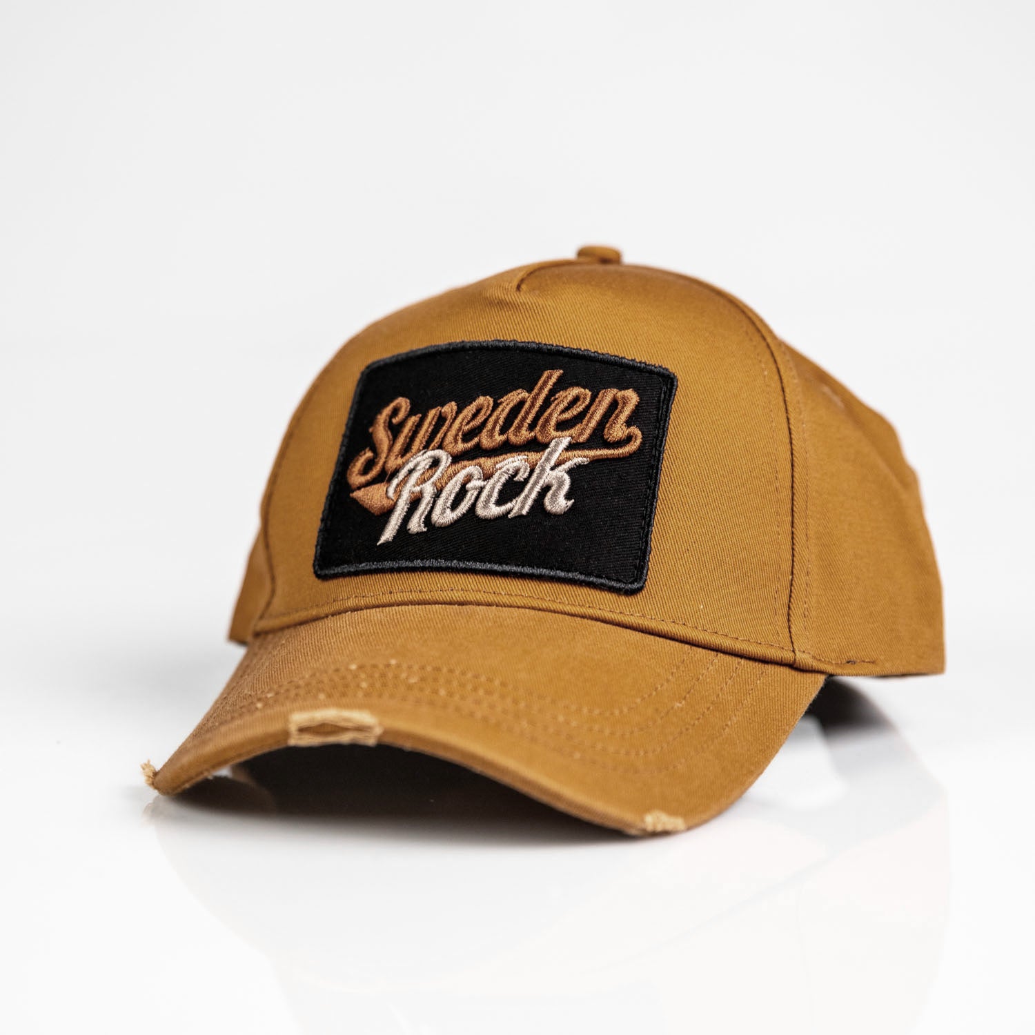Keps i brunt: En brun keps tillverkad av bomull med lätt böjd skärm och slitna detaljer, med en svart patch broderad med "Sweden Rock" i fetstil i vitt och gult på framsidan.