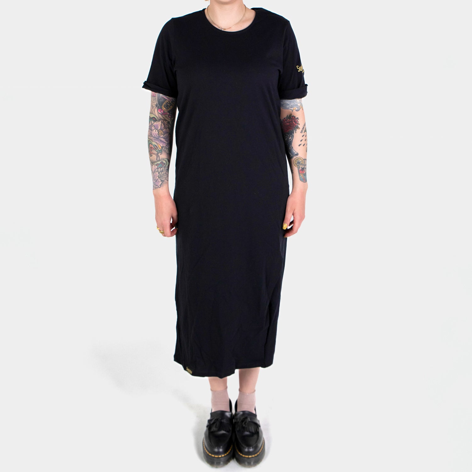 En person som bär Klänning - en svart, kortärmad T-shirtklänning i bomull - står vänd framåt och visar tatueringar på båda armarna, ljusa strumpor med svarta skor, allt mot en vit bakgrund.