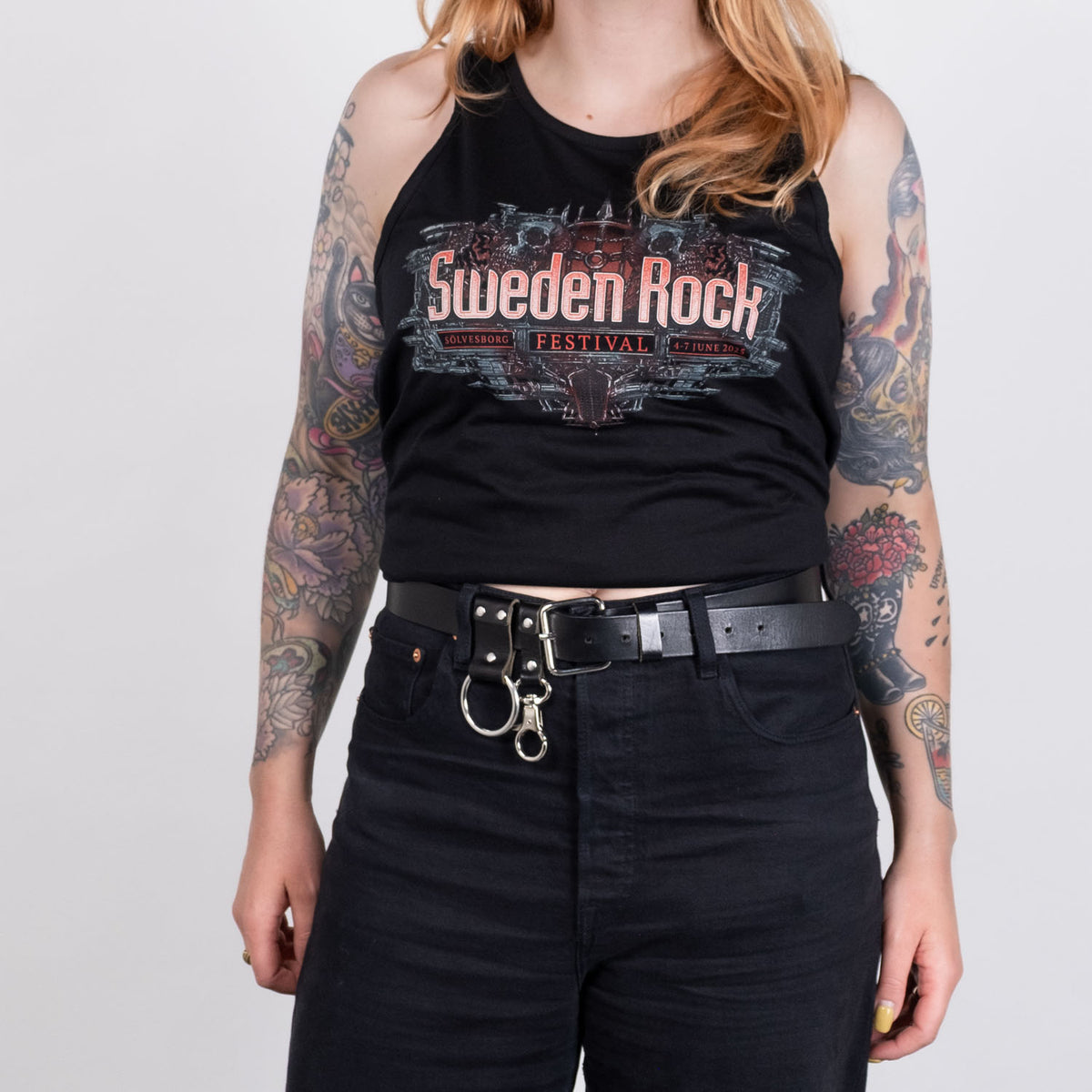 En person med långt, vågigt hår bär ett svart Festival-linne av 100% bomull, tillsammans med svarta byxor, ett svart bälte med metallringar och har färgglada tatueringar på båda armarna. Bakgrunden är enfärgad vit.