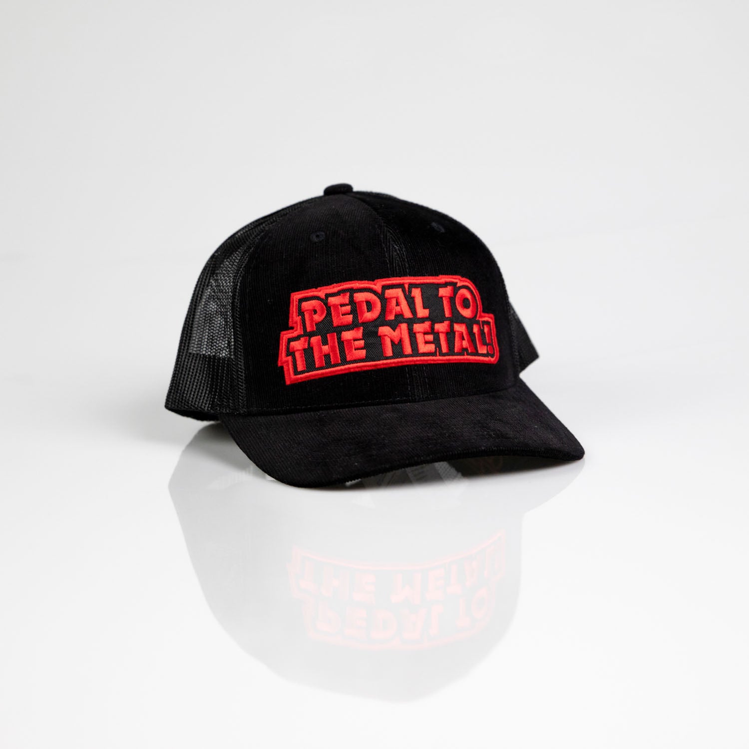 Keps Pedal to the Metal är en svart truckerkeps med mesh-sidor och en böjd skärm, med en röd och vit "PEDAL TO THE METAL" broderad patch på framsidan. Denna snygga snapback visas på en vit yta.