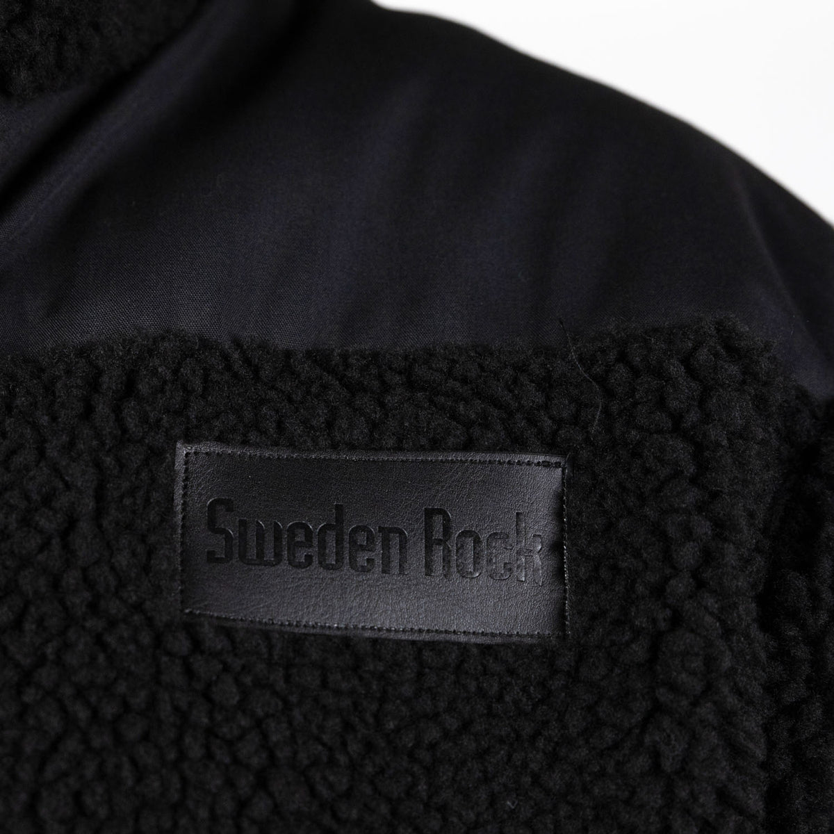 Närbild på Teddy Fleecejacket i svart, med en svart rektangulär patch präglad med "Sweden Rock". Bilden framhäver fleecens struktur och det släta tyget ovanför lappen.