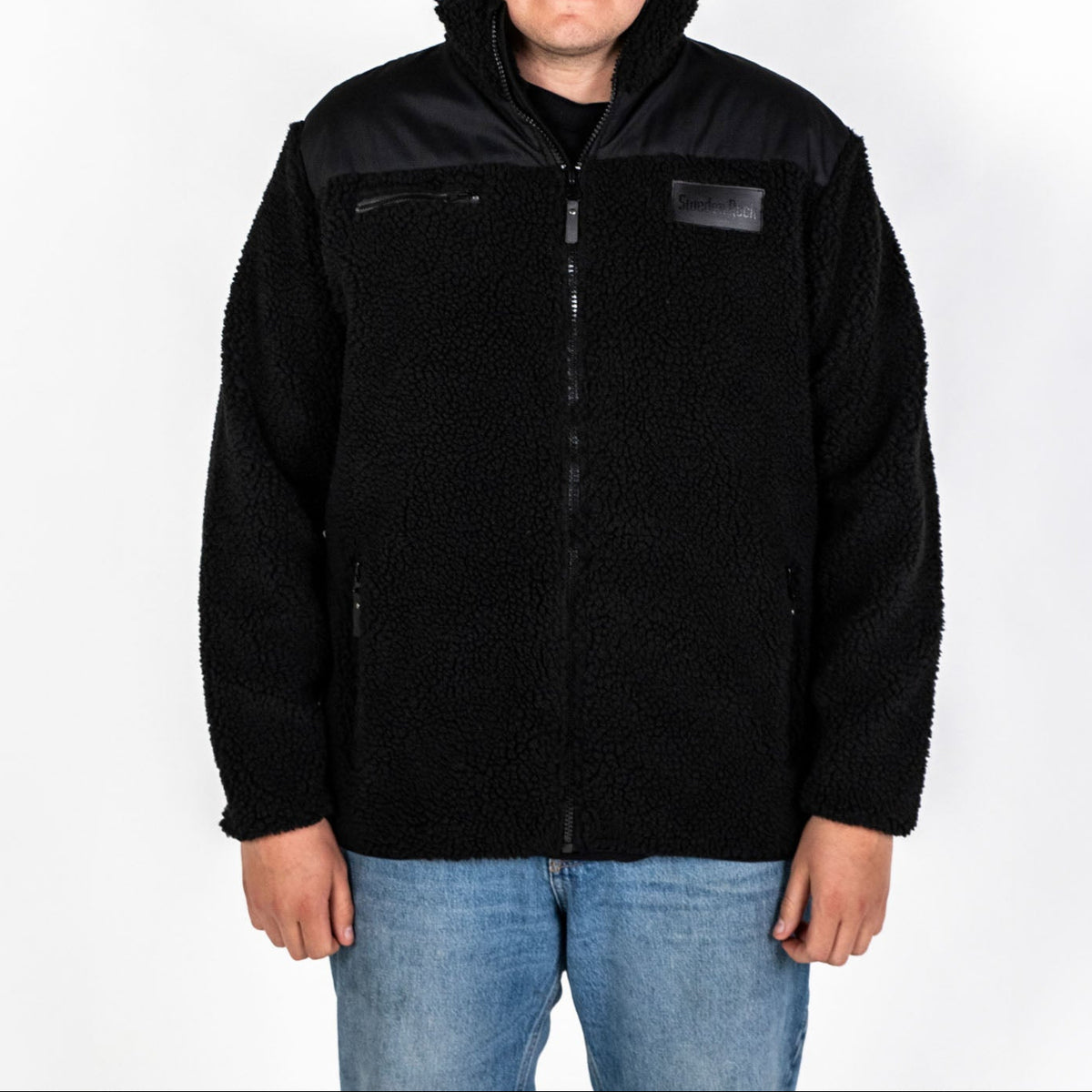 En person som bär Teddy Fleecejacket, en svart fleece med hög krage och dragkedja, tillsammans med blå jeans och mot en vit bakgrund.