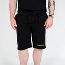 En person klädd i svarta collegeshorts med "Sweden Rock" i gult på vänster ben, en svart t-shirt, ett armbandsur och en tatuering på höger underarm.