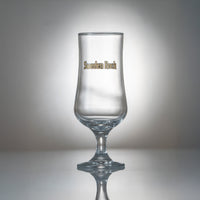 Ett klart ölglas från Ölglas 2-pack, med Sweden Rock i guld och svart på sidan, visas på en reflekterande yta mot en gråtonad bakgrund.