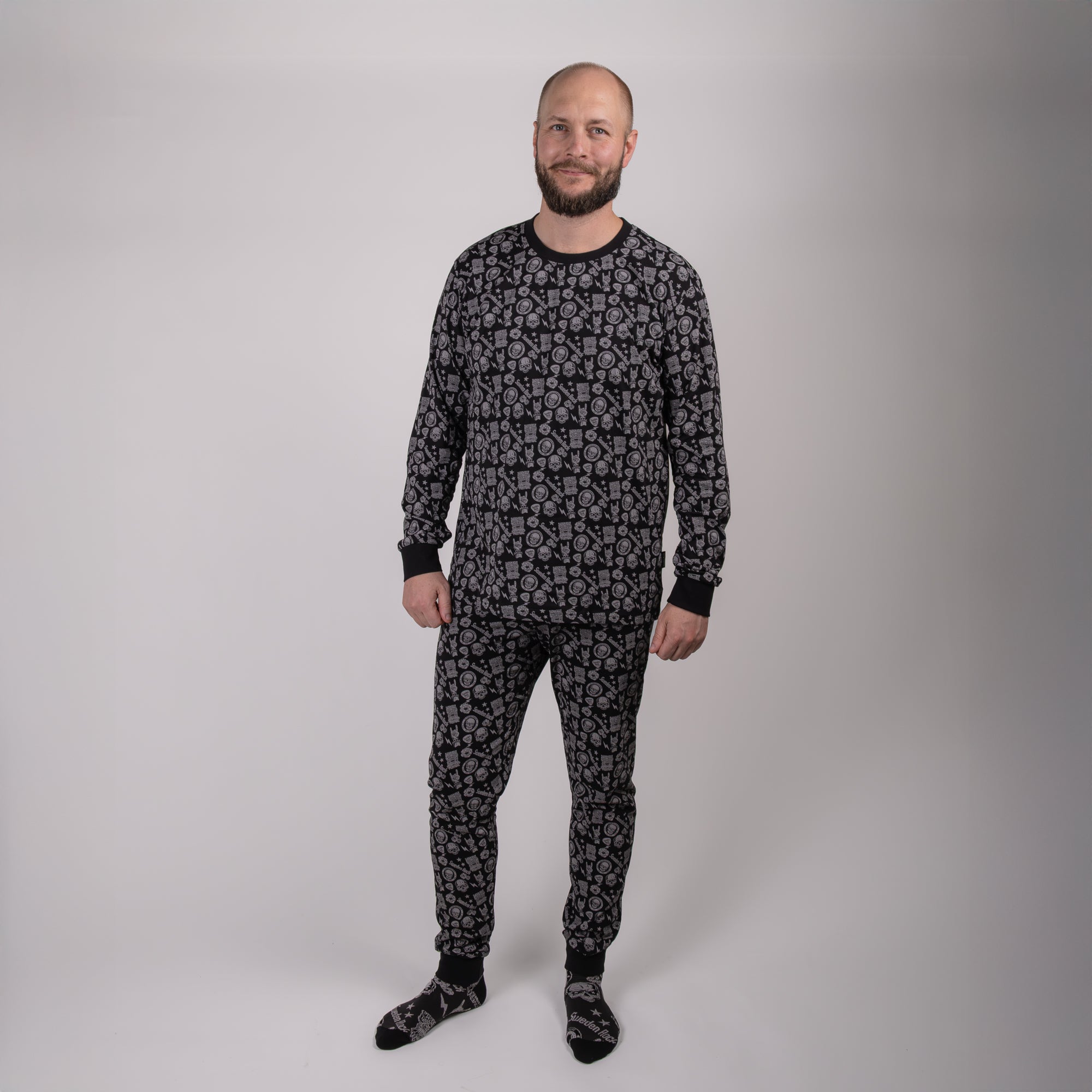 En man står i en enkel, ljus studio iklädd Pyjamas Adults i svart och grått mönster med matchande strumpor. Han är vänd framåt och ler med armarna avslappnade längs sidorna.