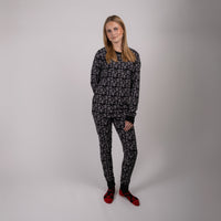 En kvinna med rakt blont hår står mot en vanlig ljus bakgrund, klädd i matchande svart och grå mönstrad pyjamas Adults och röda tofflor. Hon har händerna i fickorna och tittar in i kameran.