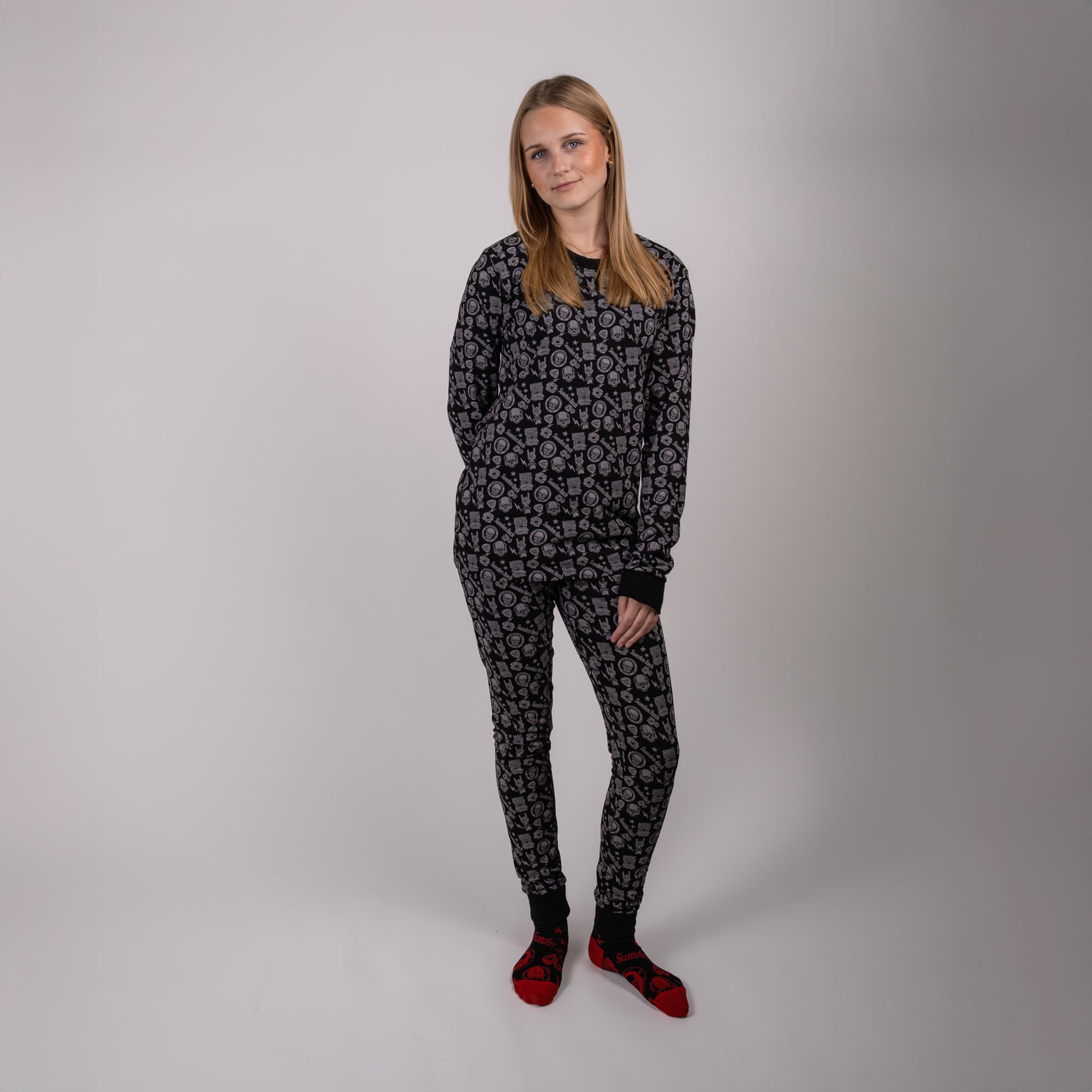 En kvinna med rakt blont hår står mot en vanlig ljus bakgrund, klädd i matchande svart och grå mönstrad pyjamas Adults och röda tofflor. Hon har händerna i fickorna och tittar in i kameran.