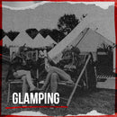 Två personer slappnar av i solstolar utanför ett Glampingtält - 2 pers uppställt på gräs. "Glamping" visas i fetstil nedan, med träd och flaggor som ger en festlig festivalboendekänsla - perfekt för glamping på Sweden Rock.