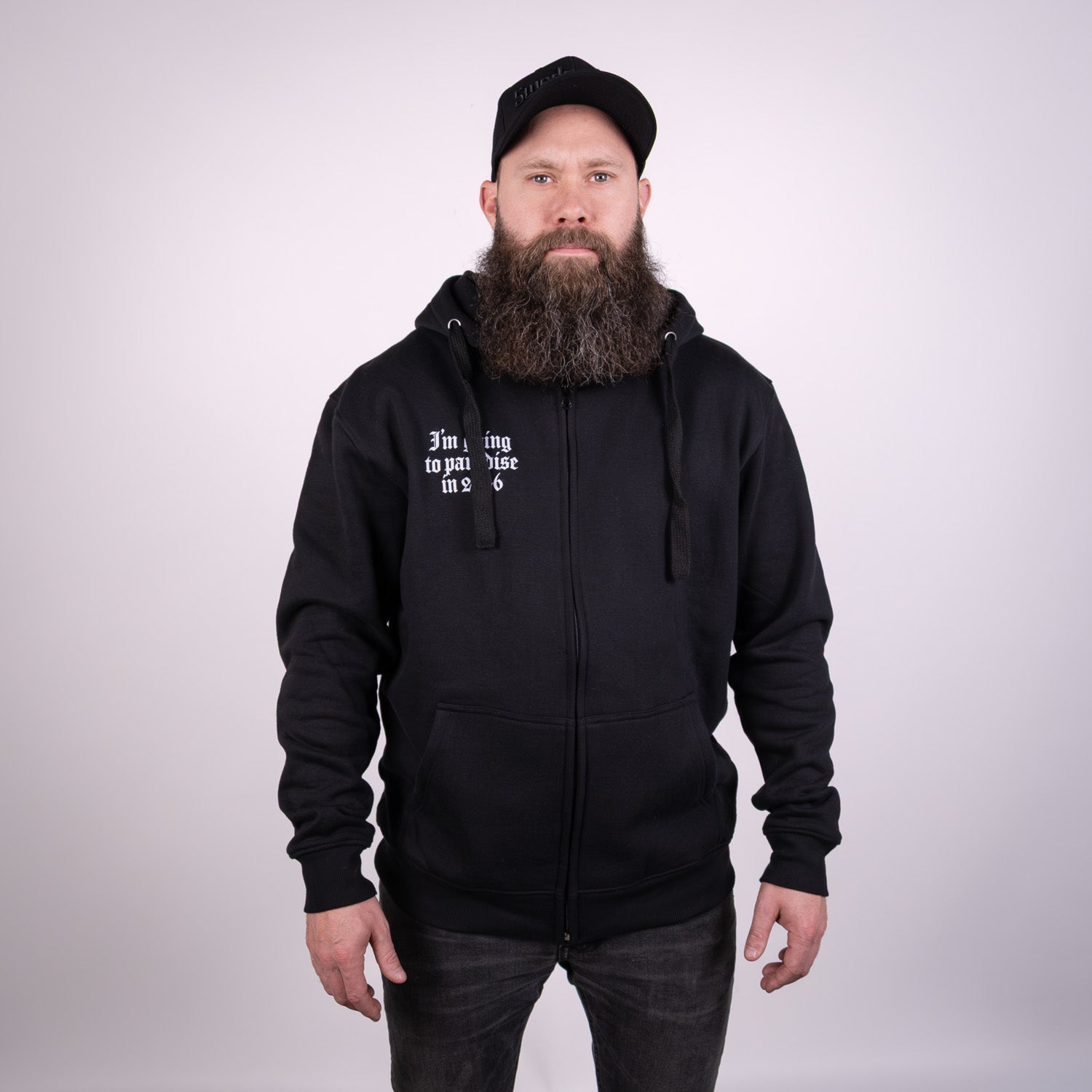 En man med långt skägg och allvarligt ansiktsuttryck vänder sig mot kameran, klädd i en svart Paradise 2026 Zip Hoodie med vit text på bröstet och en svart basebollkeps. Bakgrunden är enfärgad och ljus.