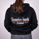 En person med långt brunt hår klädd i en svart Paradise 2026 Zip Hoodie står mot en vanlig ljus bakgrund.