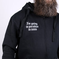 En person med långt, tjockt skägg bär Paradise 2026 Zip Hoodie, med vit text i gotisk stil på svart botten. Bakgrunden är enfärgad och ljus.